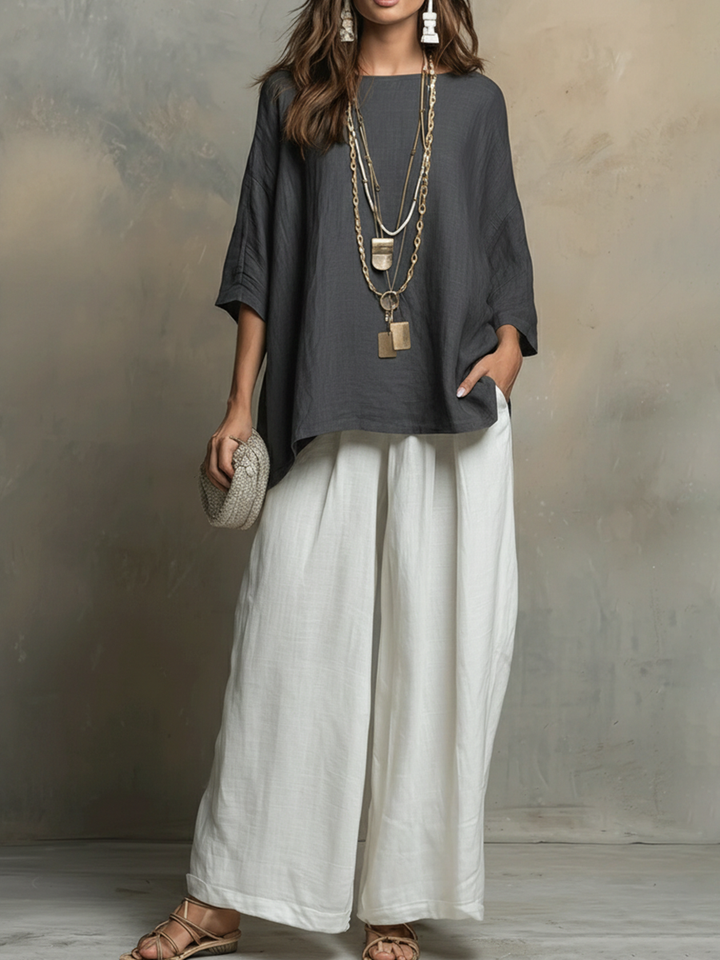 Woman in dark top with white pants, layered necklaces, holding textured clutch, studio backdrop.
