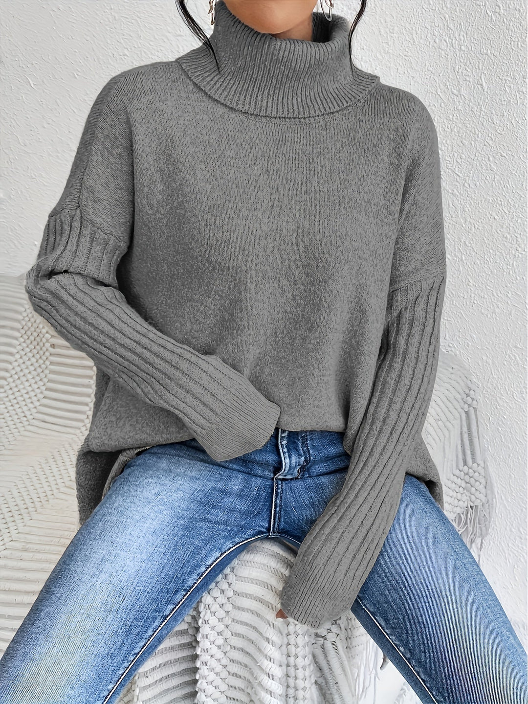 Gray sweater with turtleneck and ribbed sleeves, worn with jeans, seated casually on a textured chair indoors.