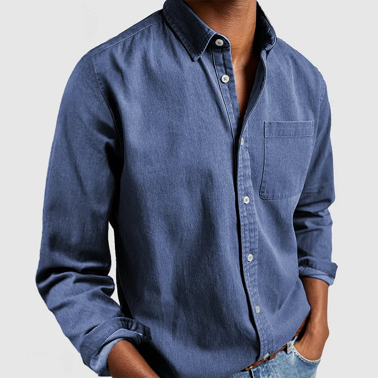 Denim blue button-up shirt with front pocket and rolled sleeves, styled with hands in pockets; gray background.