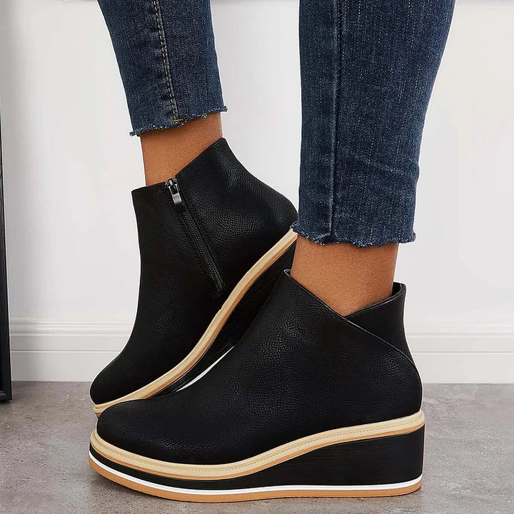 Black luxe boots with wedge sole and zipper, paired with frayed jeans, on a gray floor and white wall.