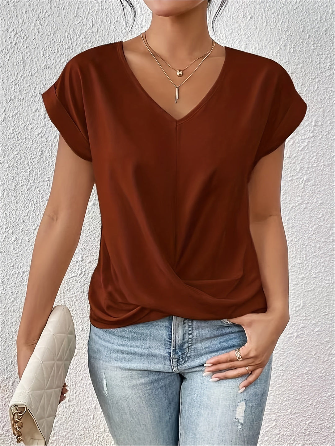 Rust V-neck T-shirt with rolled sleeves, styled with light jeans and a white quilted clutch, against a textured white wall.
