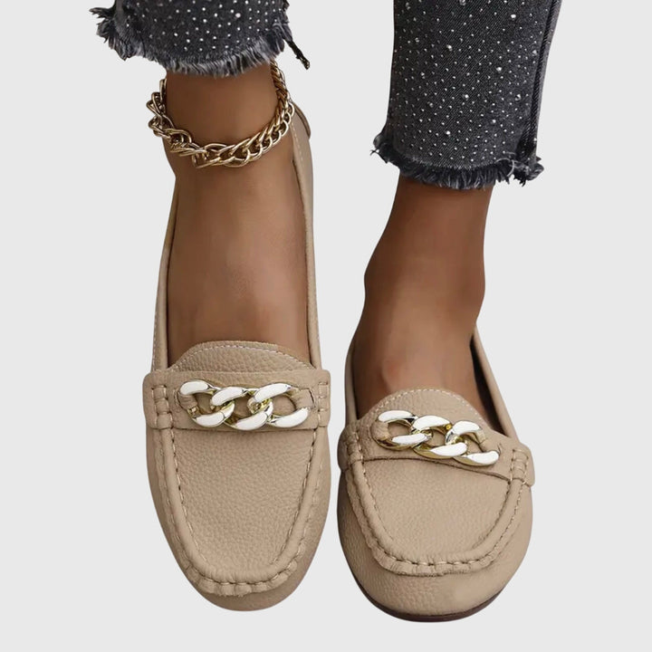 Beige orthopedic loafers with white and gold chain detail, worn with dark pants on a gray background.