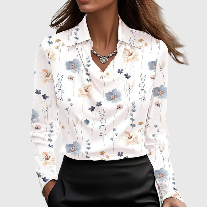 White blouse adorned with subtle pink and blue floral prints, draped neckline, long sleeves, paired with black skirt in plain setting.