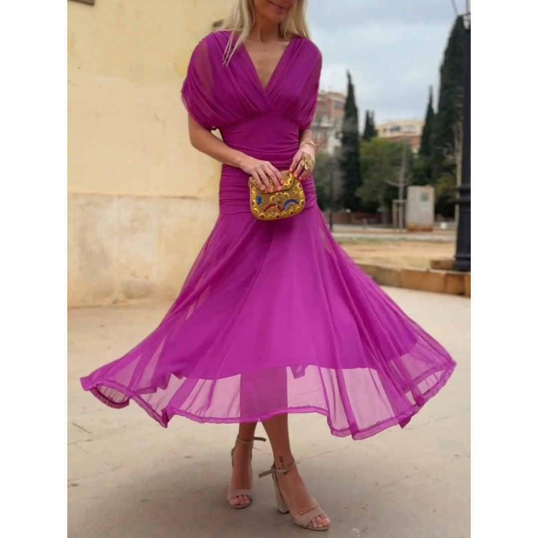 Woman in a vibrant pink mesh dress, holding a colorful clutch, standing outdoors with trees and buildings in the background.
