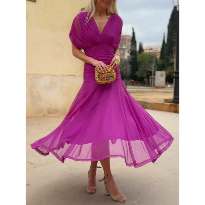 Woman in a vibrant pink mesh dress, holding a colorful clutch, standing outdoors with trees and buildings in the background.