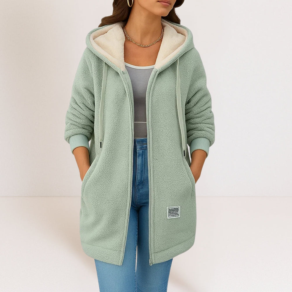 Woman in a mint green soft jacket featuring a hood, drawstrings, and pockets, styled with jeans in a minimalist studio background.