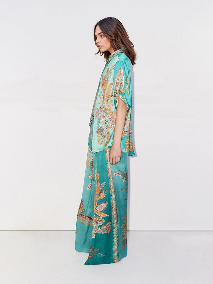 Side profile of a model in a flowing green two-piece set with paisley design, studio setting with a white backdrop.