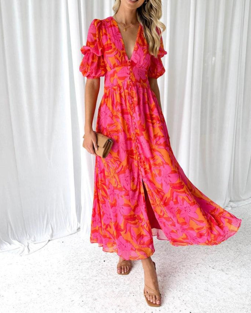 Model wearing a flowing long dress in pink and orange floral pattern, with ruffled sleeves. Holding a clutch, standing against white curtains.
