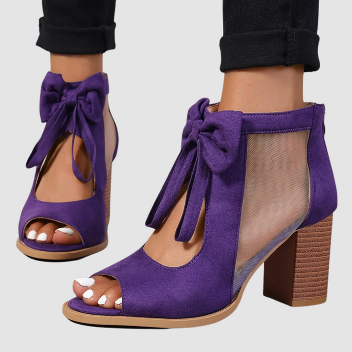 Purple heeled sandals with bow detailing, worn with black cuffed pants on a neutral background.
