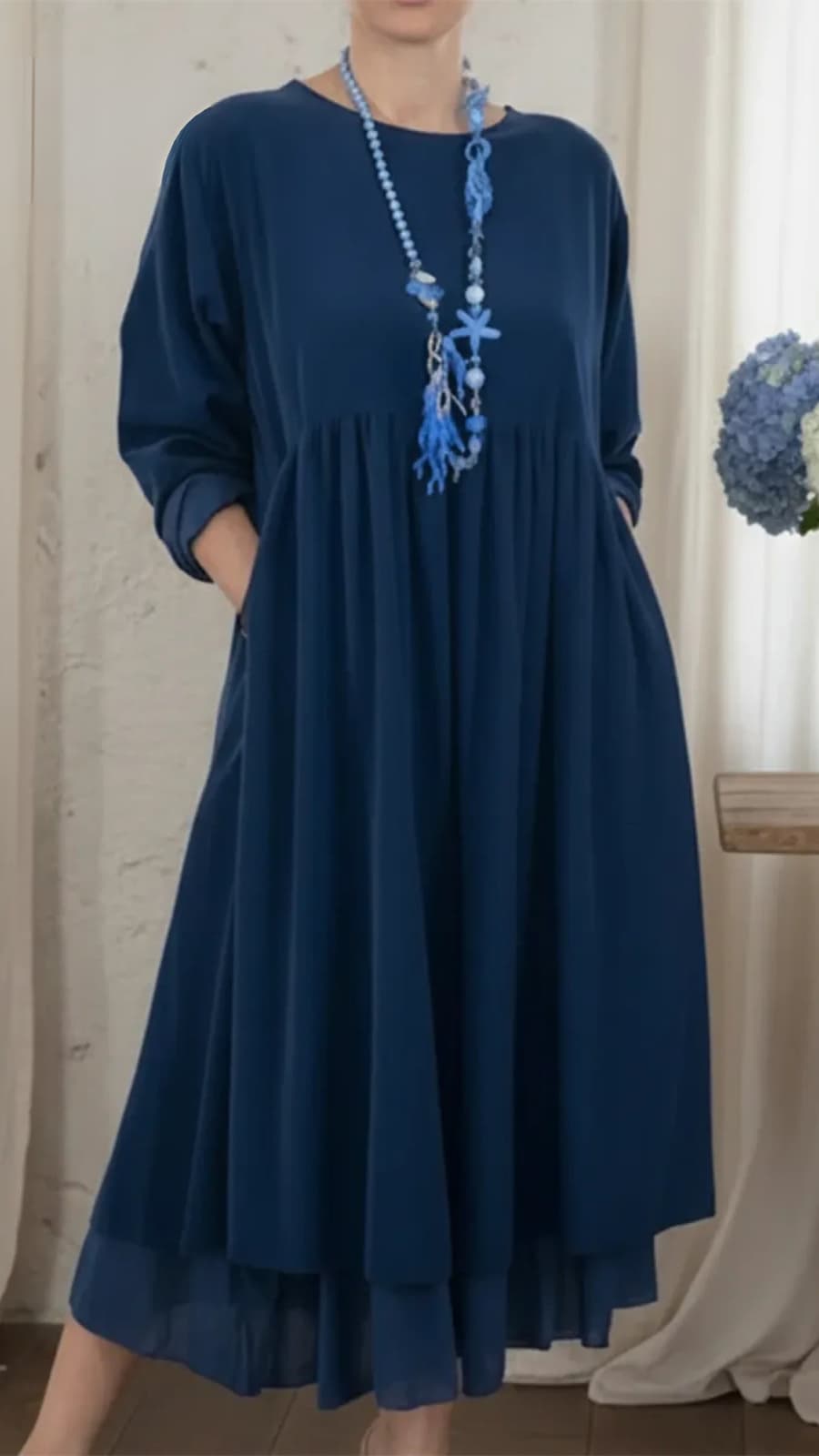 Navy blue maxi dress with long sleeves and a relaxed fit, paired with a blue beaded necklace, in an indoor setting with floral decor.