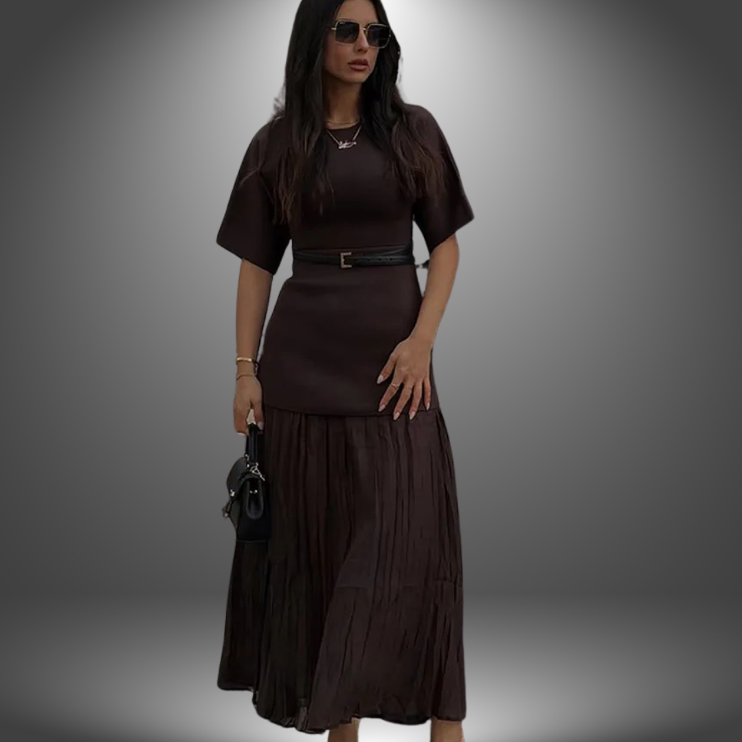 Model in dark brown handcrafted dress with pleated skirt and black belt, holding handbag against gradient studio backdrop.