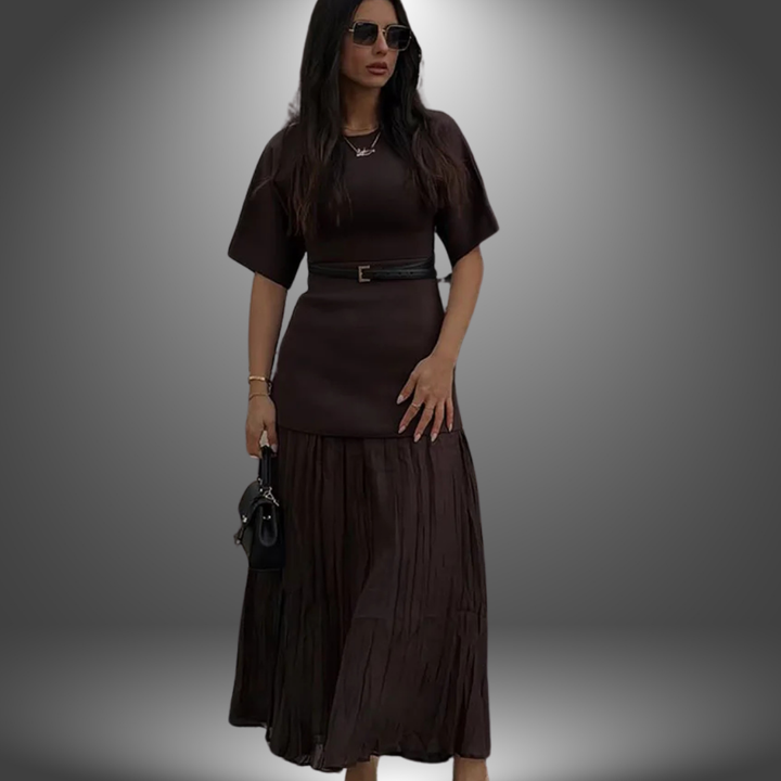 Model in dark brown handcrafted dress with pleated skirt and black belt, holding handbag against gradient studio backdrop.