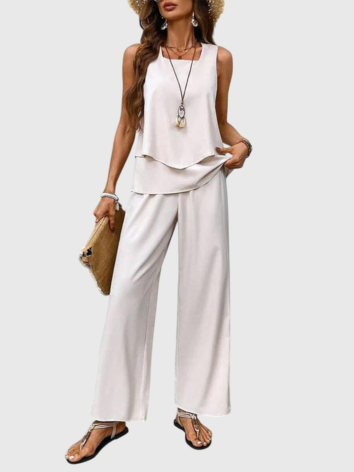 Model in white sleeveless top with flowing hem and white wide-leg pants. Neutral backdrop.