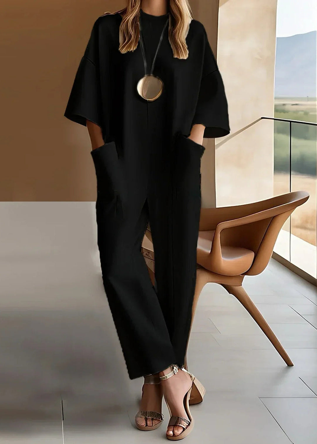 Woman in a black short-sleeve jumpsuit with side pockets, posing in a stylish room with brown chair and large window view.