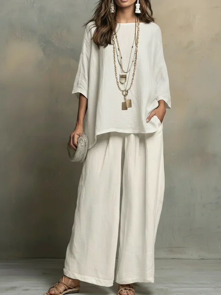 Woman in flowing white two-piece set with layered necklaces, holding textured clutch, studio backdrop.
