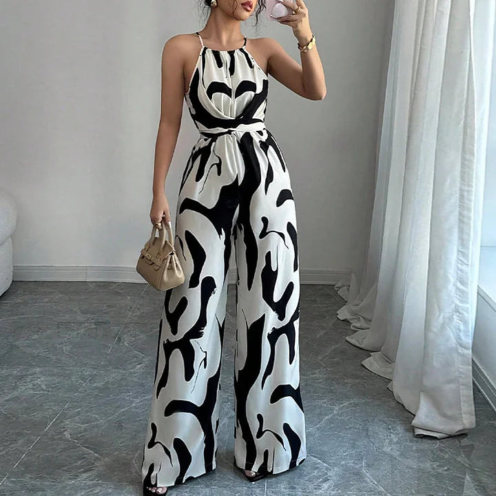 Black and white patterned backless jumpsuit with halter neck, worn by a model in a bright room with sheer curtains.