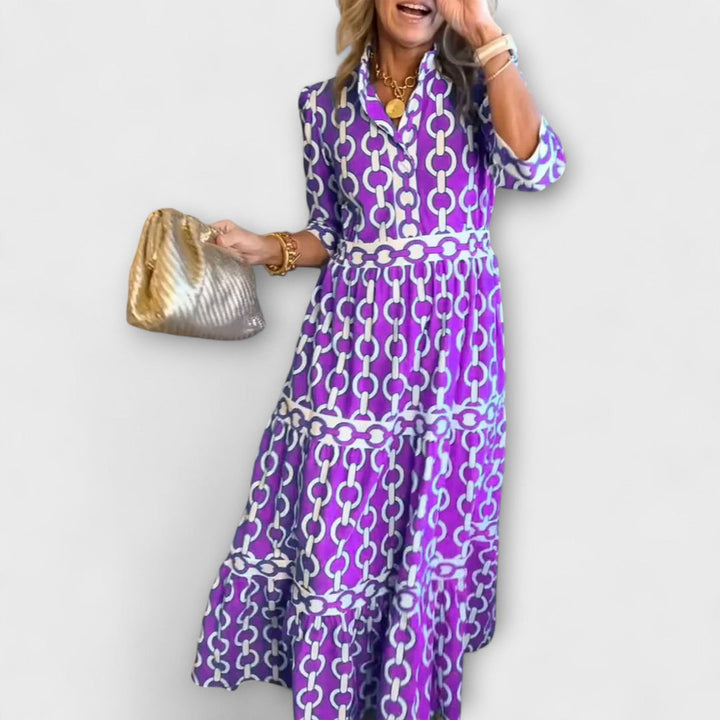 Woman in purple and white chain print midi dress holding a gold bag against a plain background.