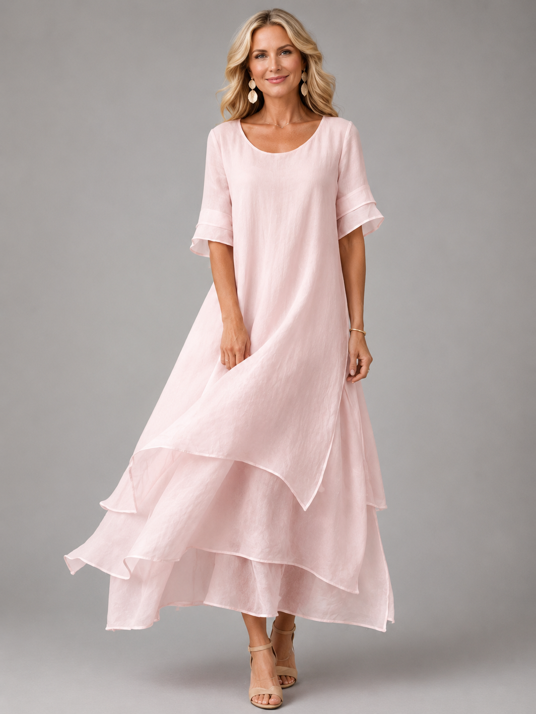 Woman in soft pink flowing maxi dress with layered hem, standing in studio, smiling.