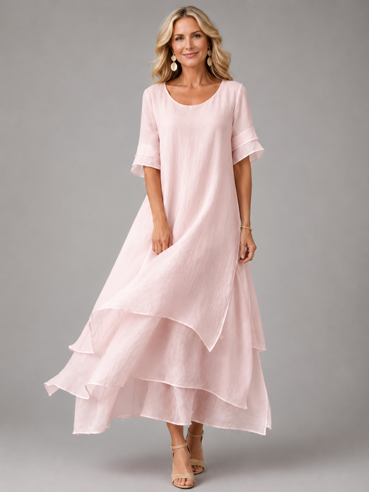 Woman in soft pink flowing maxi dress with layered hem, standing in studio, smiling.