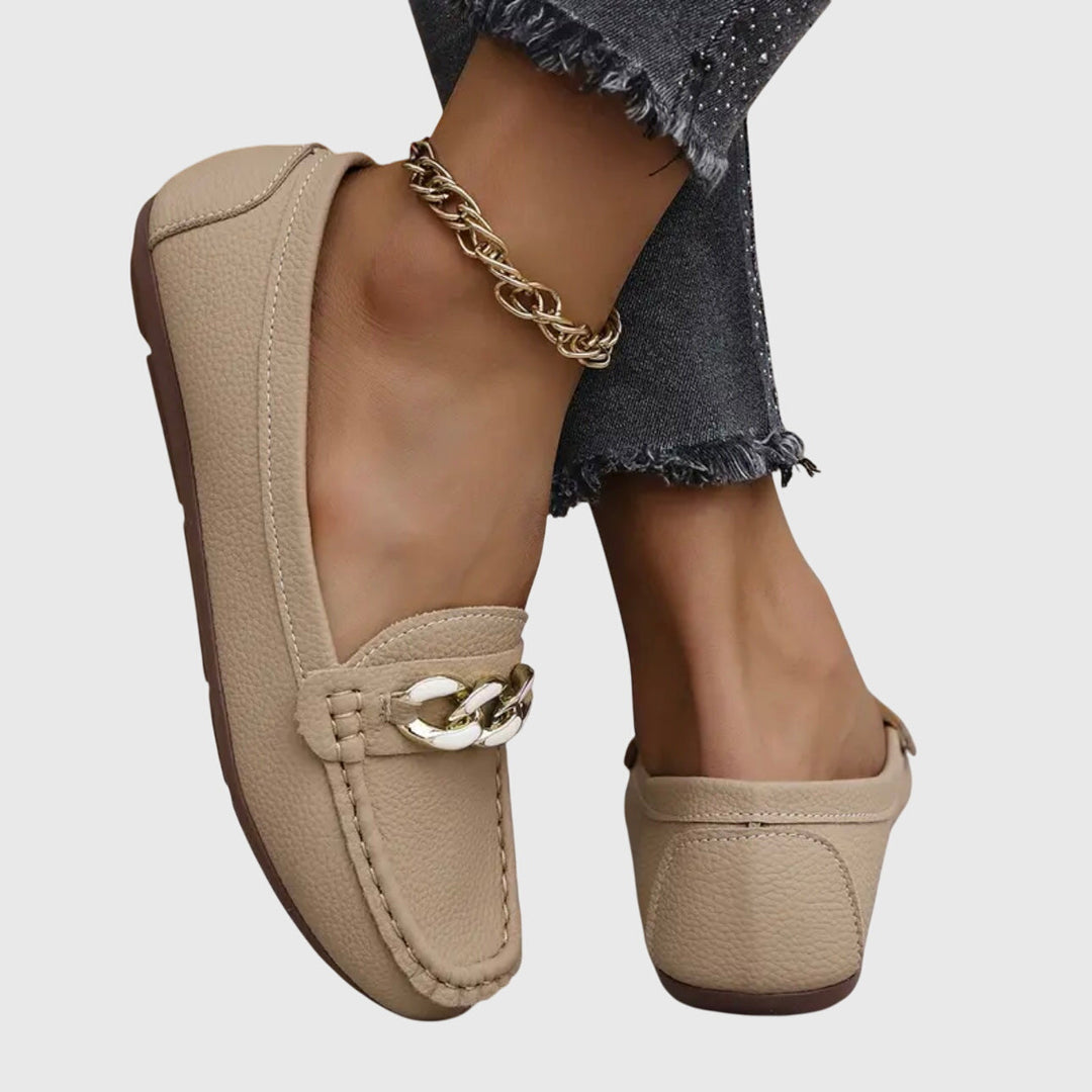 Back view of beige orthopedic loafers with gold and white chain detail, worn with dark pants on a gray background.