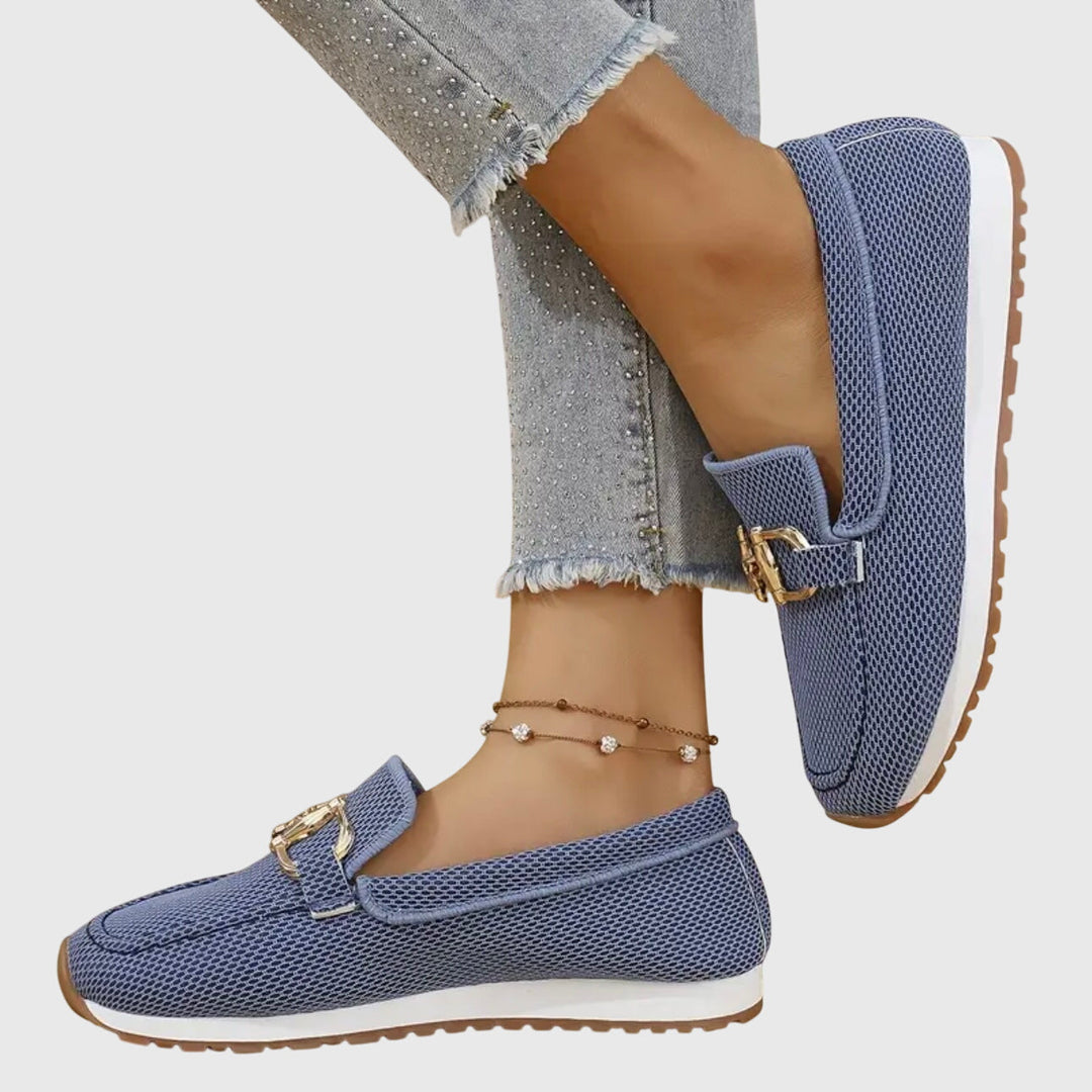 Side view of blue orthopedic loafers with gold details worn with frayed jeans and anklet on a light background.