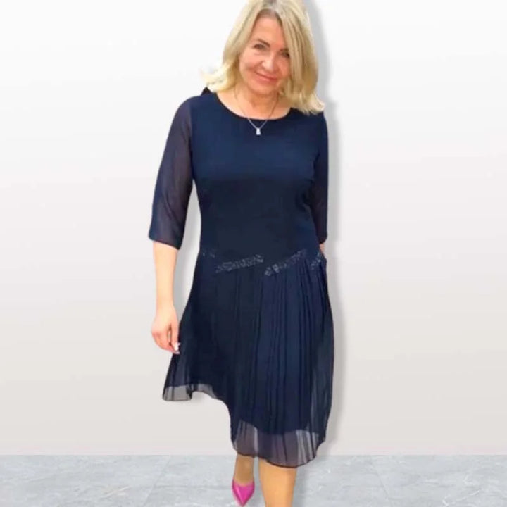 Woman in navy elegant midi dress with 3/4 sleeves, pleated skirt, standing on gray tile floor in a studio setting.