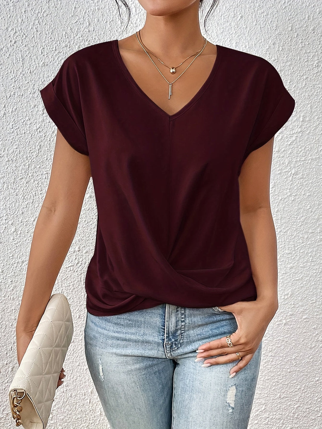 Maroon V-neck T-shirt with rolled sleeves, styled with light jeans and a white quilted clutch, against a textured white wall.
