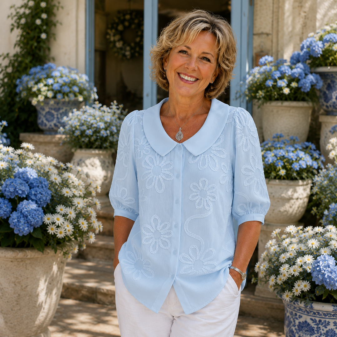 Woman in a light blue floral blouse poses in garden setting with blue and white flowers.