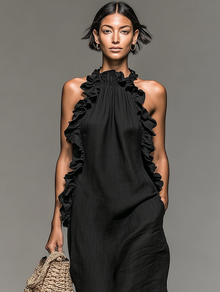 Front view of black maxi dress with ruffled neckline and side pockets, model holds a woven bag against gray background.