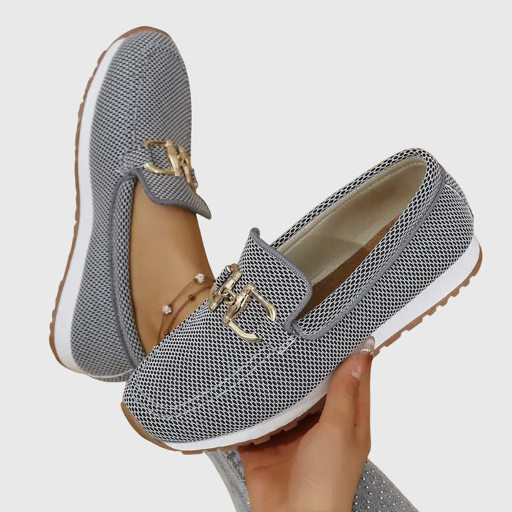 Gray orthopedic loafers with gold embellishments held in hand, paired with anklet and frayed jeans on a light background.