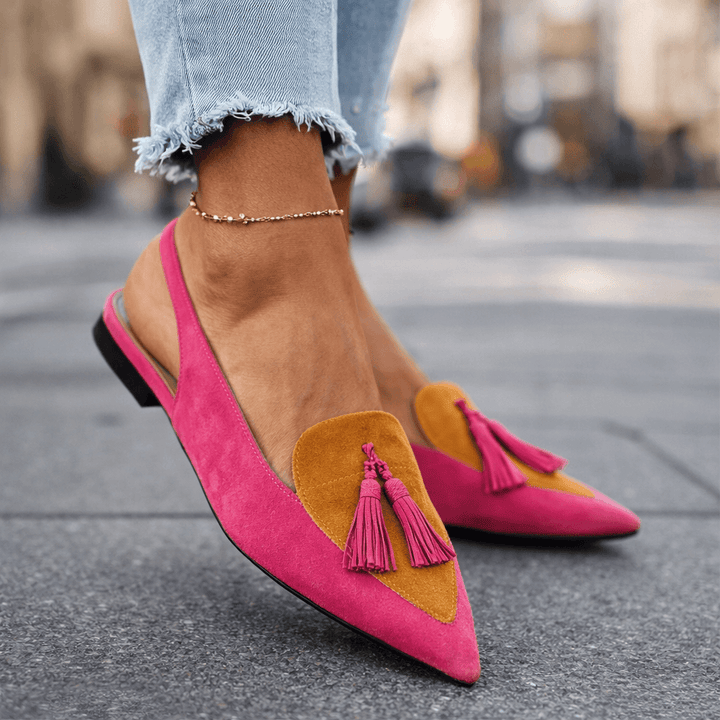 Pink sandals with mustard accents and tassels, worn with jeans, cityscape background.