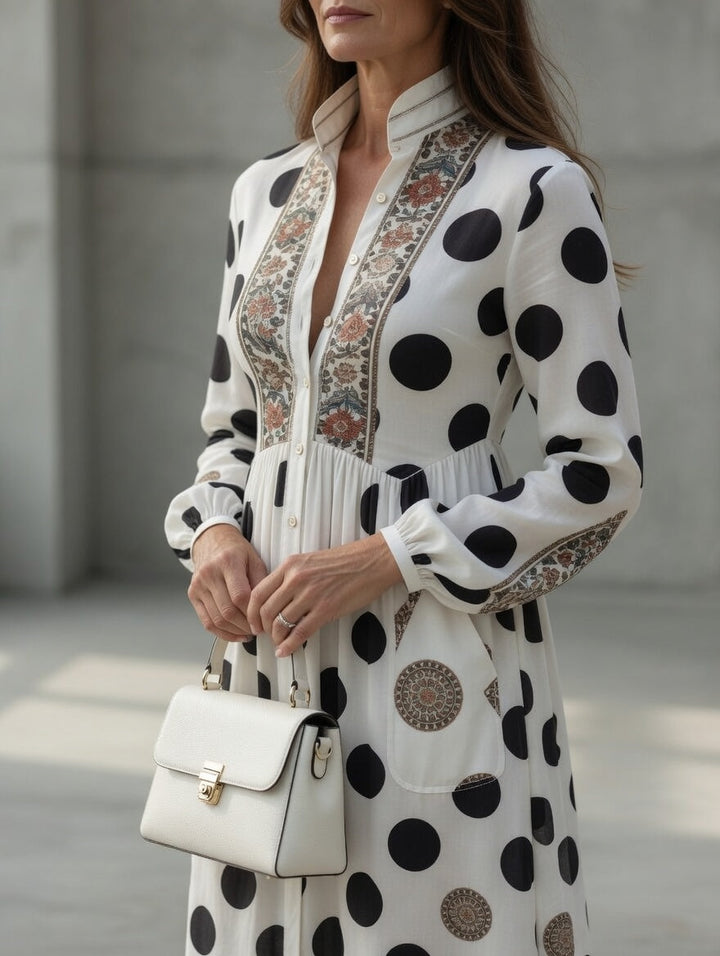 Close-up of elegant maxi dress featuring polka dots and detailed patterns, accessorized with a white handbag in a minimalist space.