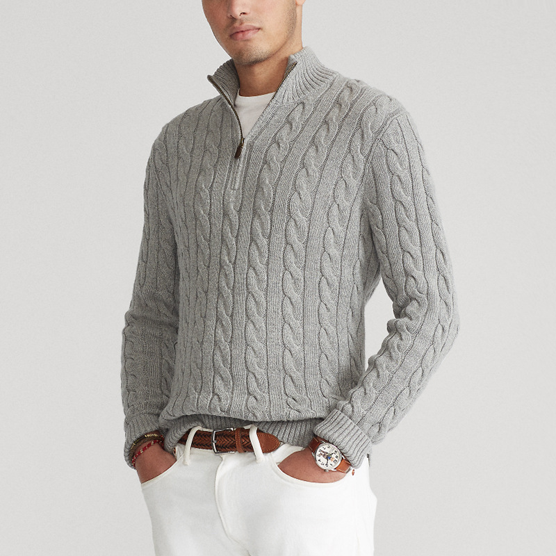 Person wearing a gray cable-knit half-zip sweater over a white shirt, hands in pockets, in a studio setting.