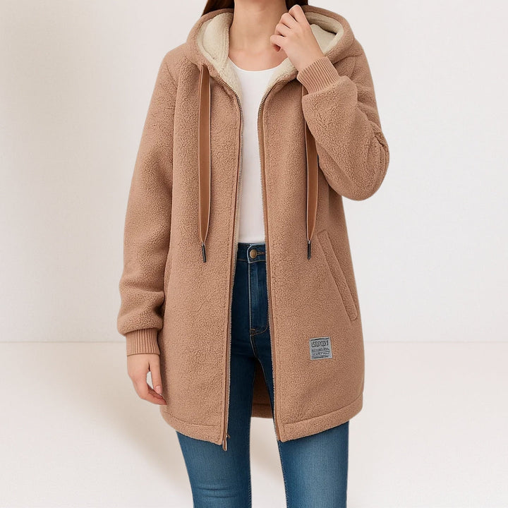 Person in a beige soft jacket with a hood and drawstrings, matched with jeans against a plain studio backdrop.