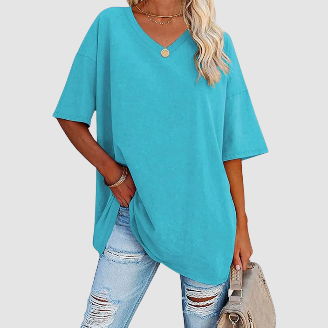Woman in a turquoise V-neck T-shirt with distressed jeans, holding a beige bag, studio background.