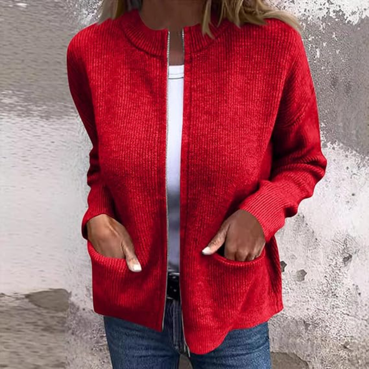 Red cozy cardigan worn over a white t-shirt with hands in pockets. Textured wall background.