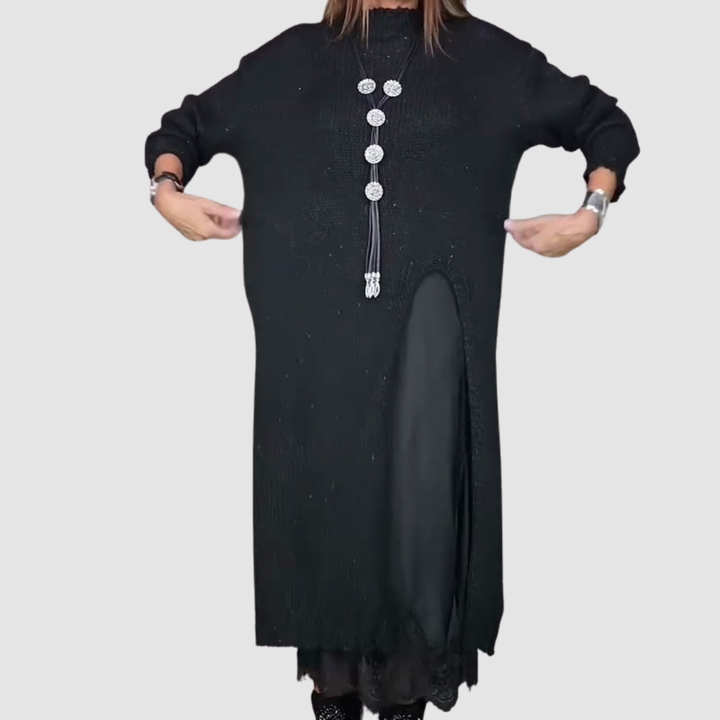 Woman stretching sides of a black dress, showing layered design and necklace, against a plain gray backdrop.