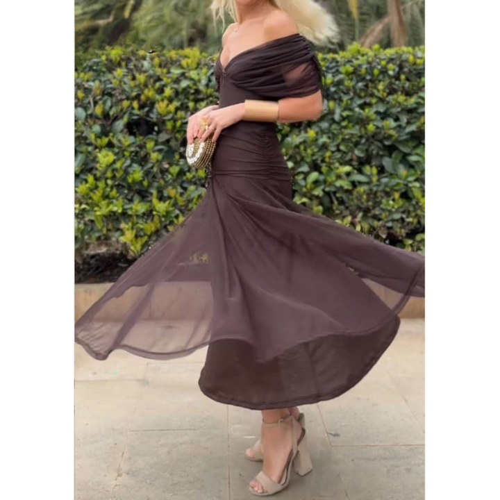 Woman in an off-shoulder brown mesh dress, standing on a walkway with greenery in the background.