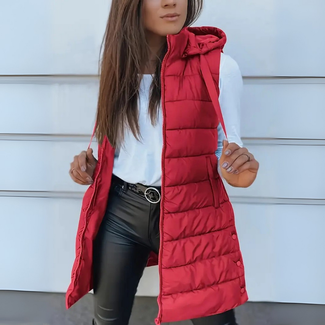 Red sleeveless puffer vest with hood worn over white top and black pants, outdoor setting.