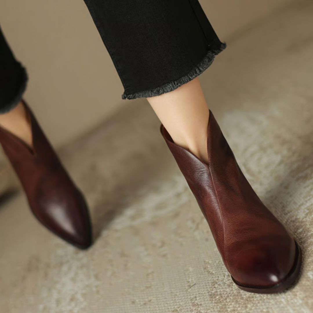 Top view of brown ankle boots with pointed toes, paired with black frayed pants on a textured surface.