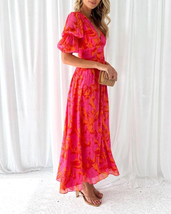 Side view of a model in a flowing long dress with pink and orange floral design, holding a clutch. Standing in front of white drapes.