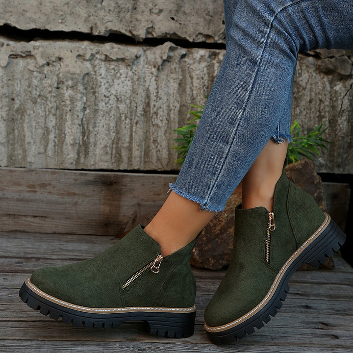 Green ankle boots with zipper, worn with frayed jeans against a textured concrete wall.