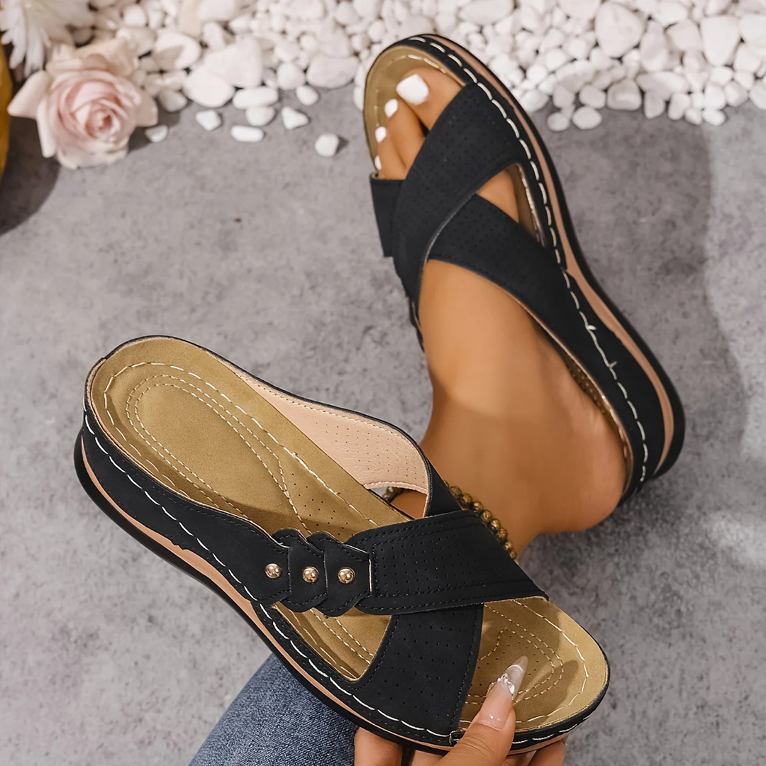 Black orthopedic wedge sandals displayed on feet, with a pink rose and white stones on a textured surface.