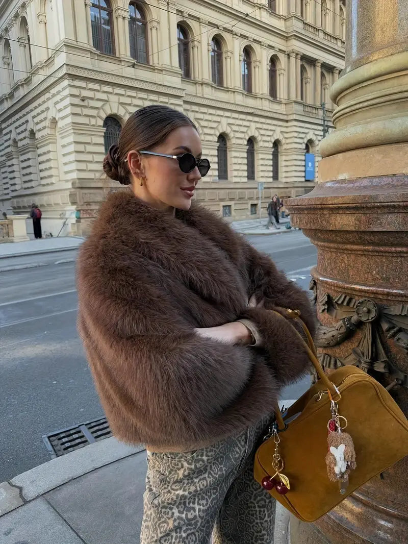 Individual wearing a stylish brown fur jacket with patterned pants, holding a mustard-colored bag, standing near an ornate building column.