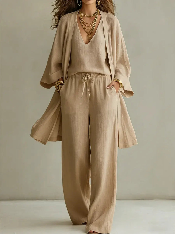 Beige three-piece relaxed homewear set with v-neck top, drawstring pants, and flowy overcoat in a studio setting.