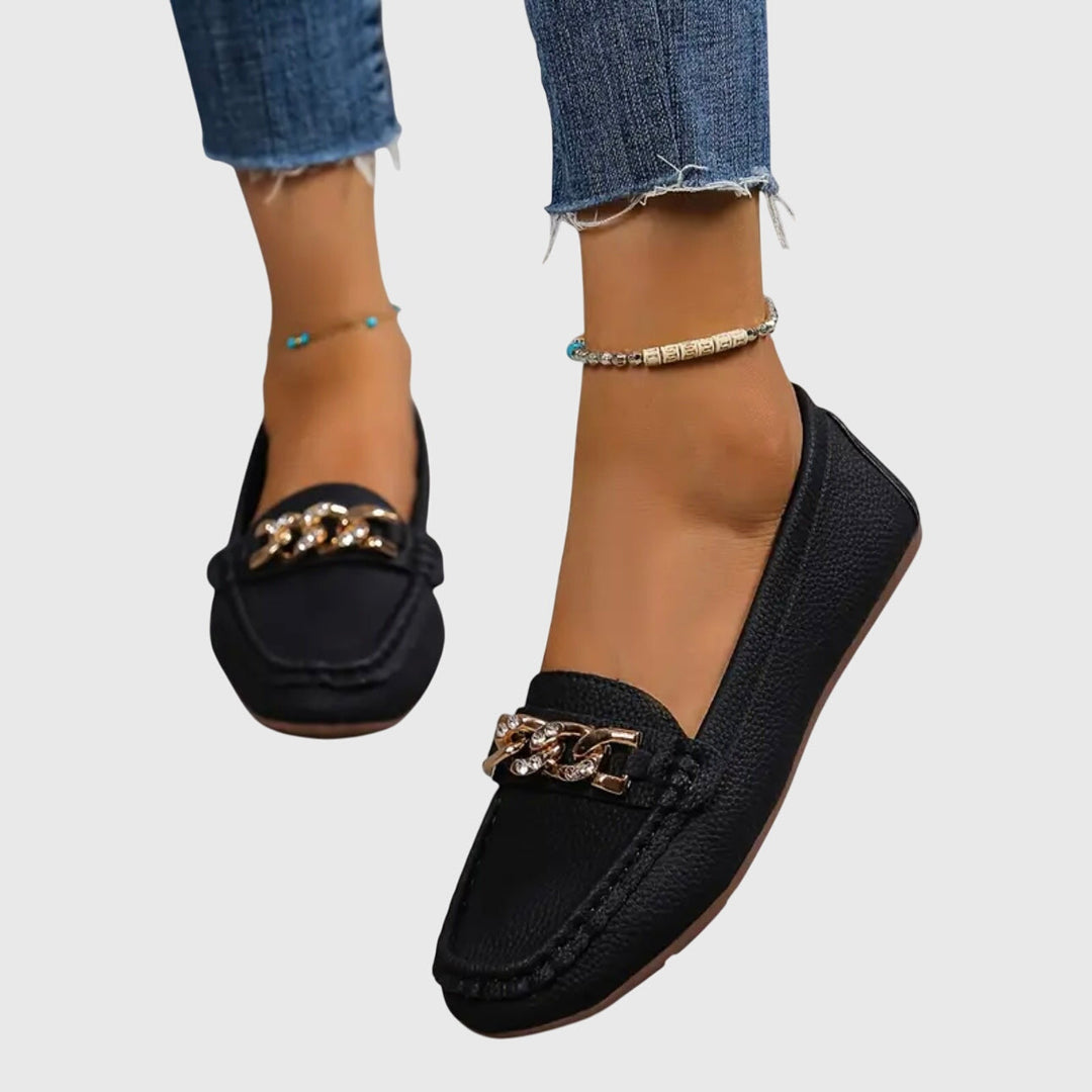 Black orthopedic loafers with gold chain detail, worn with blue jeans on a gray background.