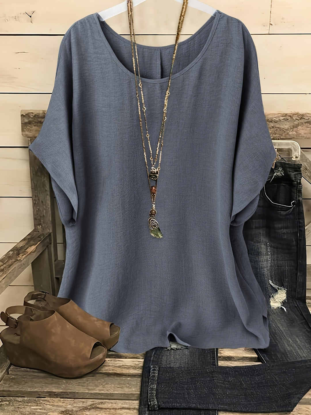 Gray loose summer top on hanger with necklace, sandals, and jeans on rustic bench.