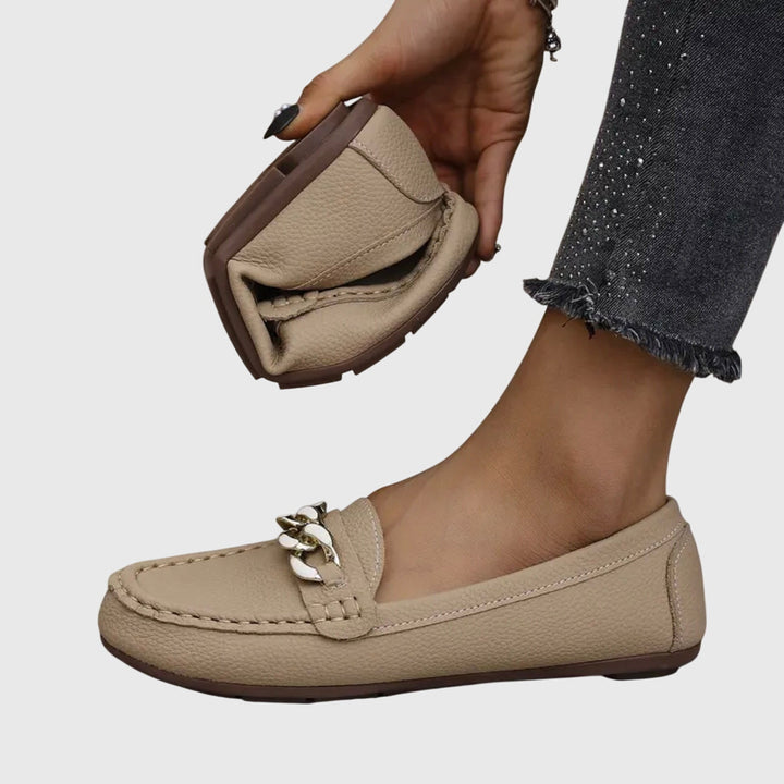 Beige orthopedic loafers with a flexible sole, one being folded, with dark pants on a gray background.