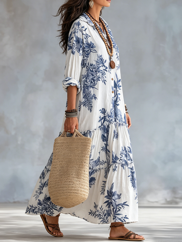 Side profile of a white maxi dress with blue floral designs, accessorized with necklaces and bracelets, carrying a woven bag.