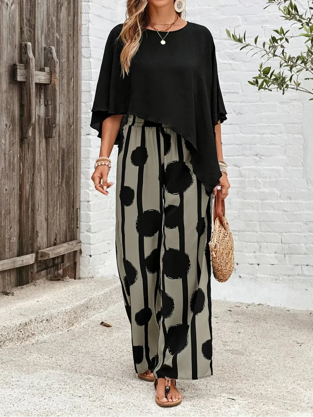Front view of a woman in a black top and abstract patterned grey pants, holding a woven bag, outdoors with a white brick background.